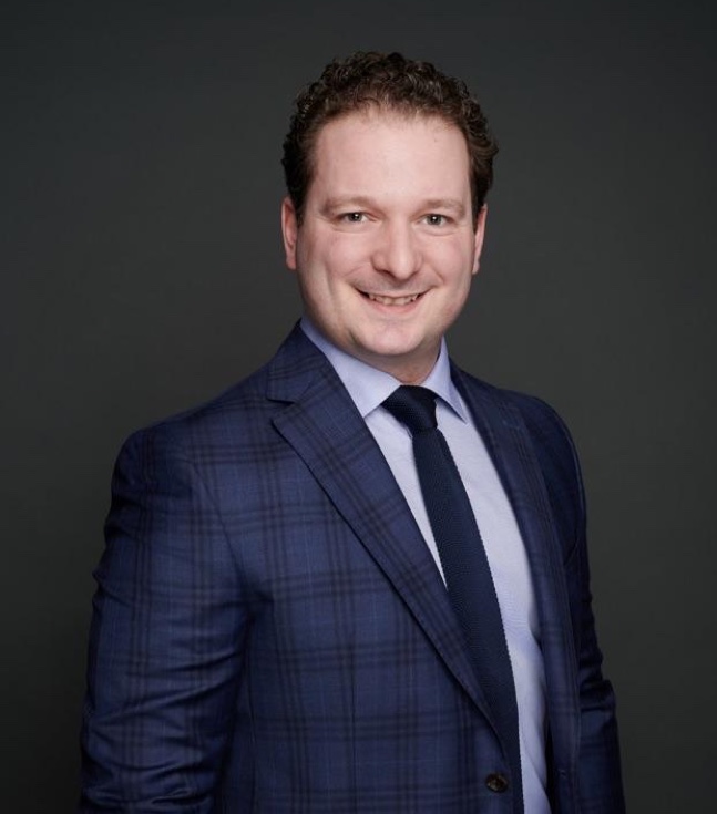 Portrait professionnel d’un avocat criminel souriant, vêtu d’un costume bleu à carreaux, offrant une image de confiance et de compétence.